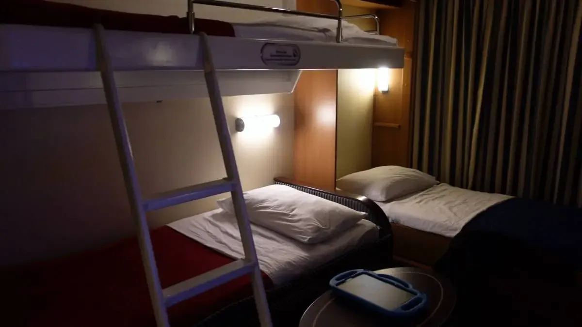 A cozy cabin room features a bunk bed with a ladder leading to the upper bunk, and a single bed below, both neatly made. Soft lighting illuminates the wooden walls and striped curtains, creating a warm atmosphere.