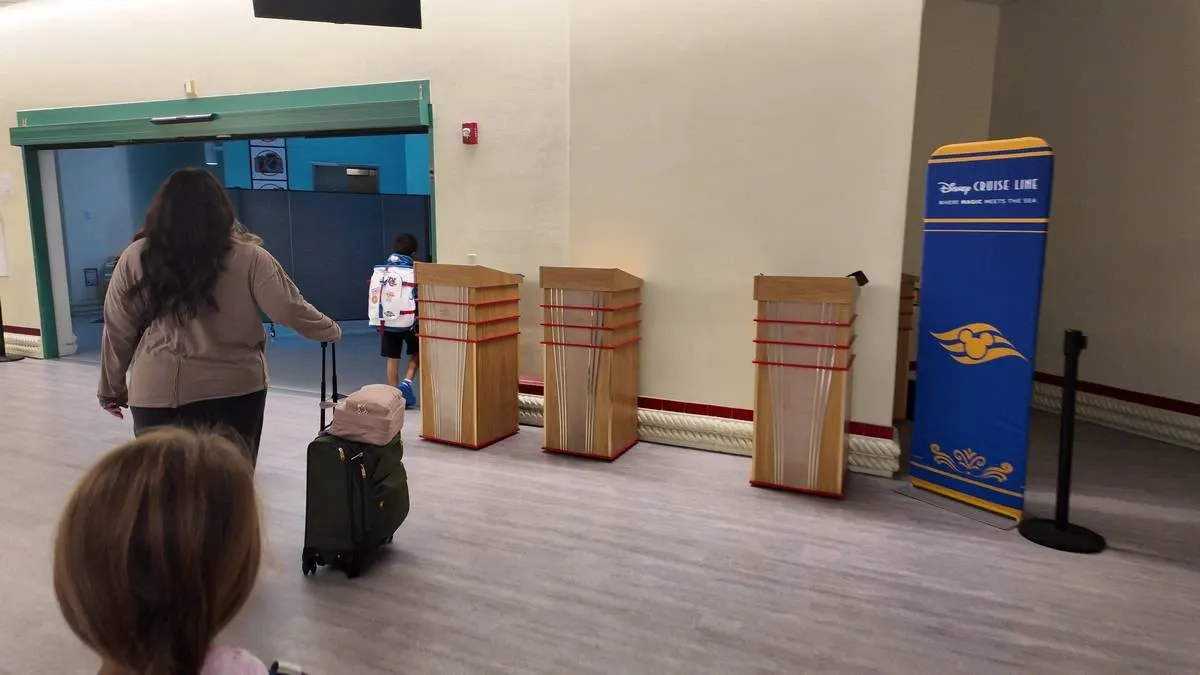 A woman with a suitcase and a child walk towards a green doorway in a hallway. A Disney Cruise Line sign is visible on the right, next to some wooden podiums.