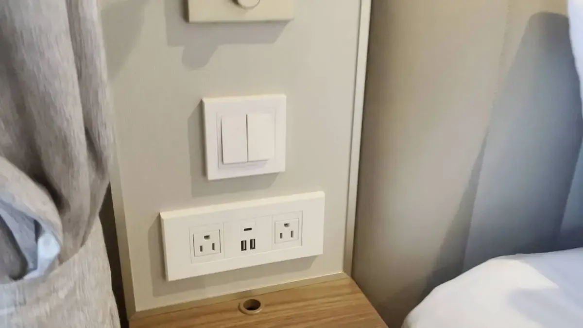 A wall panel featuring a light switch and two power outlets with USB ports is set above a wooden nightstand, partially covered by a curtain on the left and a bed on the right.