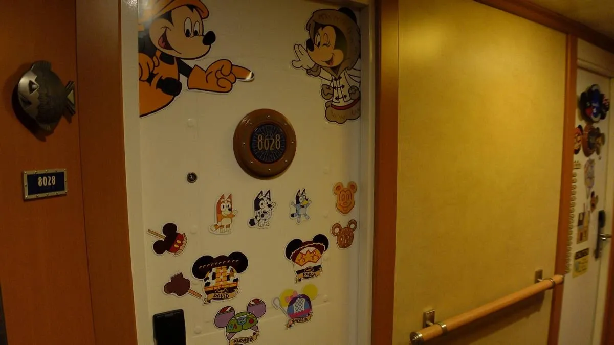 A cruise ship cabin door decorated with colorful cartoon magnets, including characters resembling Mickey Mouse and others. The door is white, and the decorations add a playful touch to the hallway setting.