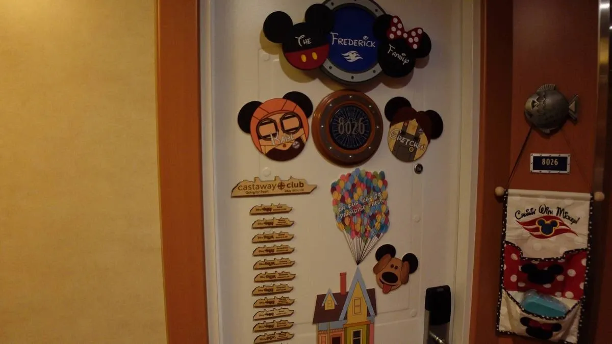 A decorated cruise cabin door features various Disney-themed magnets, including Mickey Mouse ears and a design inspired by the movie "Up" with colorful balloons. The door has a circular room number plaque, and a hanging sign welcomes guests. A fish decoration and a fabric pocket organizer with a Disney Cruise Line logo are also visible.