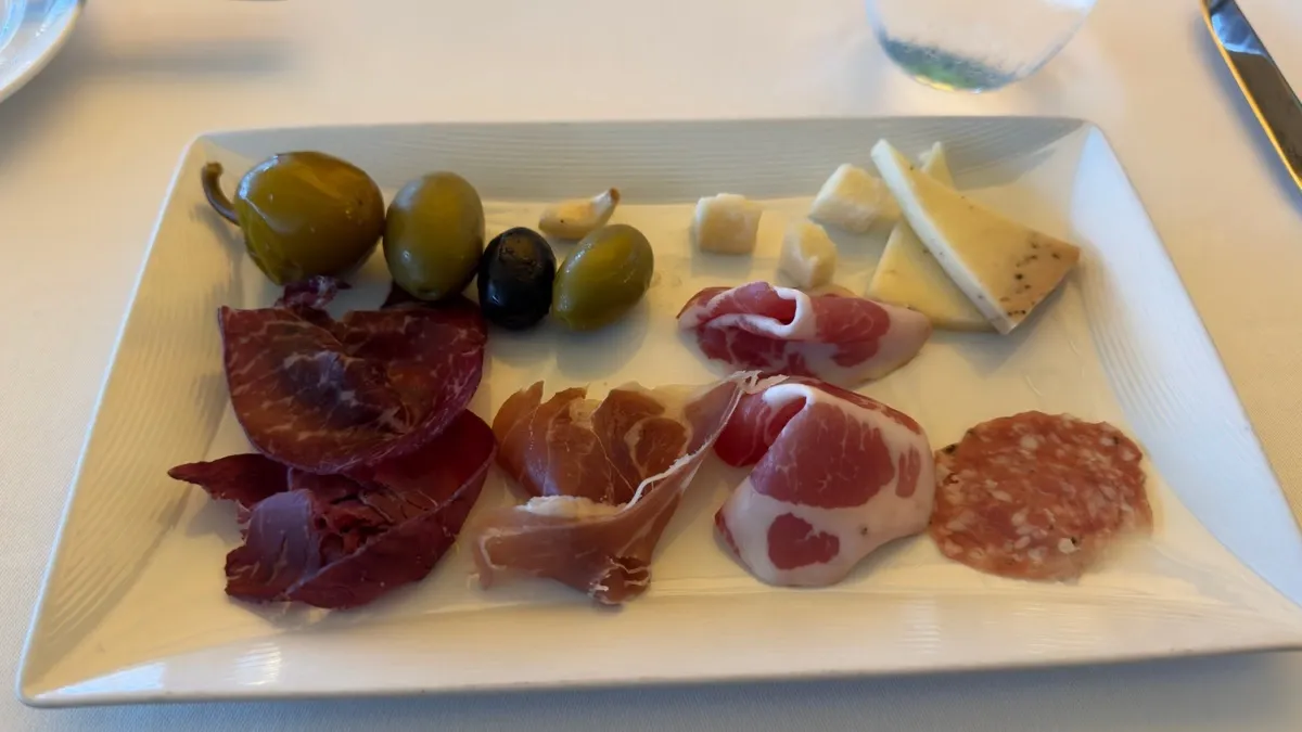A rectangular plate features a selection of charcuterie and cheese. There are slices of cured meats, including salami and prosciutto, alongside olives, caper berries, and small cubes and wedges of cheese. The arrangement is neatly presented on a white tablecloth.