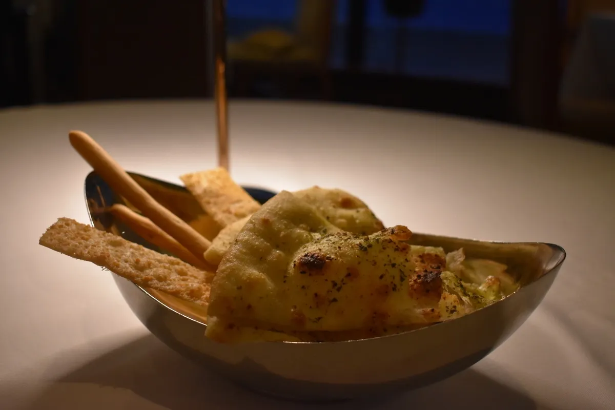 A metal bowl on a table holds a variety of crispy breadsticks and seasoned flatbreads, with a warm, inviting glow highlighting their textures.