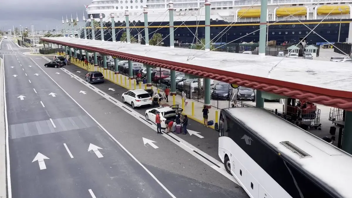 A busy cruise terminal with a large ship docked in the background. Several cars and a bus are parked along the covered drop-off area, where people are unloading luggage. The road is marked with white arrows, directing traffic flow, and the sky appears overcast.