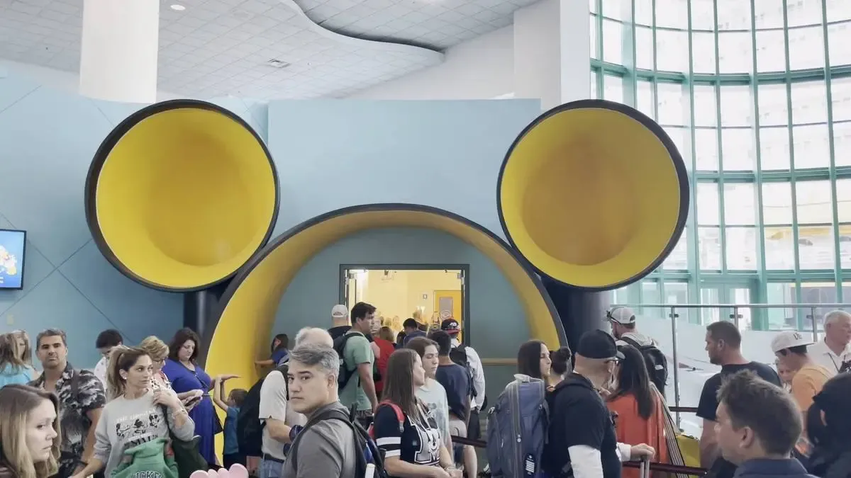 A group of people, some with luggage, gather in front of a large entrance shaped like Mickey Mouse ears, featuring prominent yellow interiors, inside a spacious, sunlit atrium with green-framed windows.
