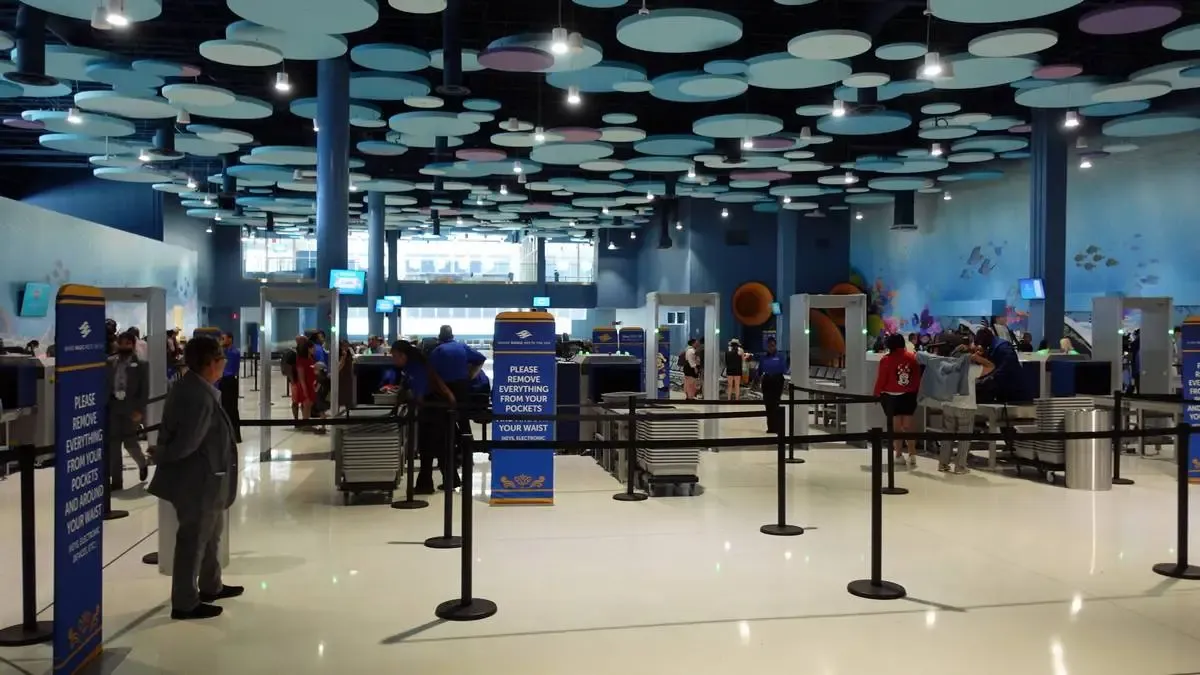 A spacious security checkpoint area with people lining up at metal detectors, surrounded by blue and white circular ceiling decorations. Signs instruct travelers to remove items from their pockets, and staff assist at the screening stations. The atmosphere is bright and organized.
