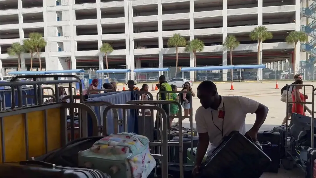 A person in a white shirt is handling luggage in an area with metal barriers, while a line of people waits in the background. The setting is outdoors, with a multi-story parking garage and palm trees visible across the street.
