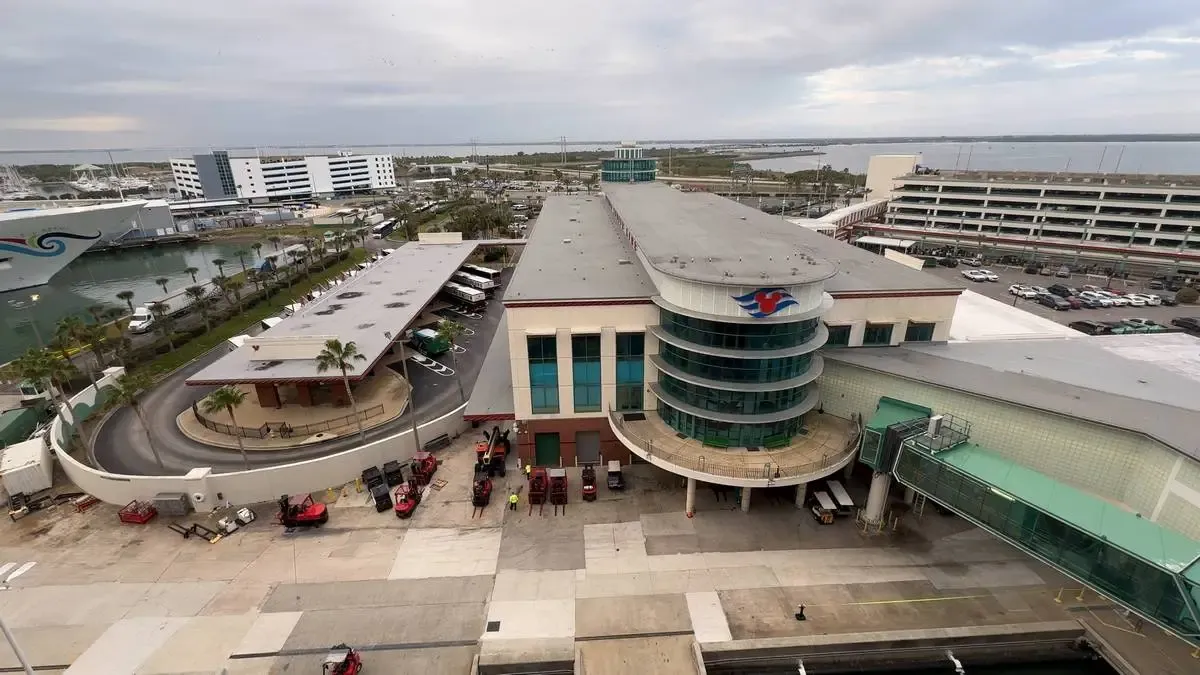 An aerial view of a cruise terminal with a large, modern building featuring a circular glass facade. Several small vehicles are parked outside, and a covered walkway connects the terminal to a ship docked nearby. The surrounding area includes parking structures and a scenic waterfront.