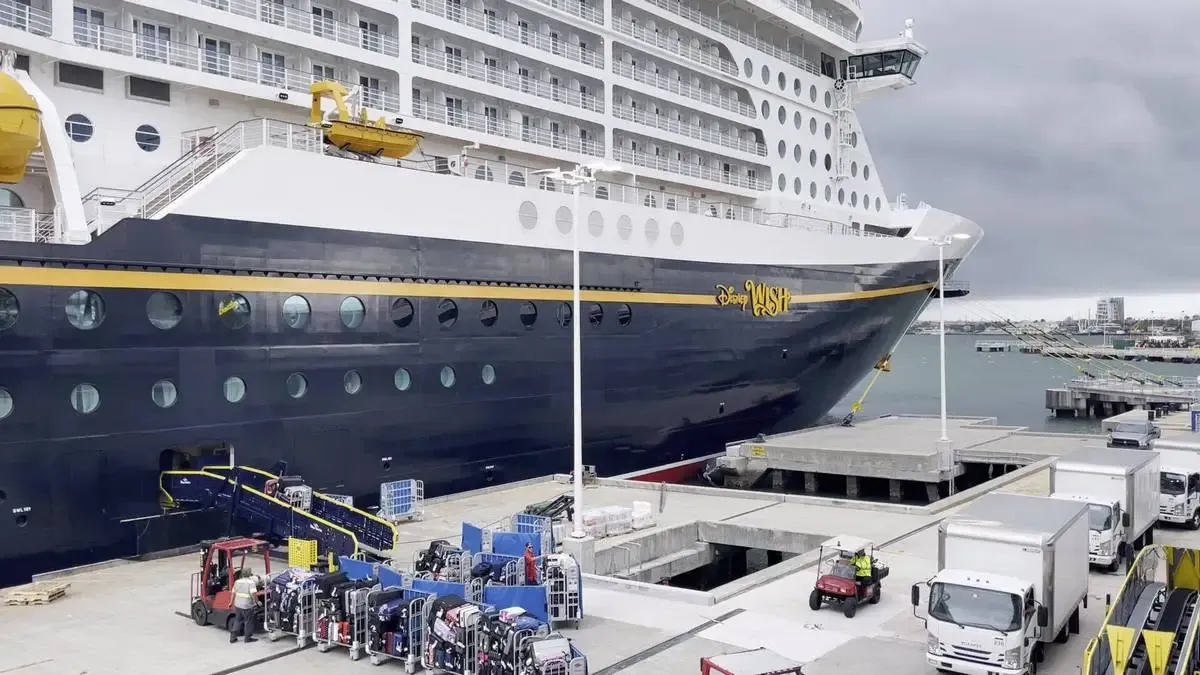 A large cruise ship with the name "Disney Wish" is docked at a port. The ship has a sleek navy and white exterior with multiple decks and rows of circular windows. Several trucks and workers are visible on the dock, handling supplies and equipment under a cloudy sky.