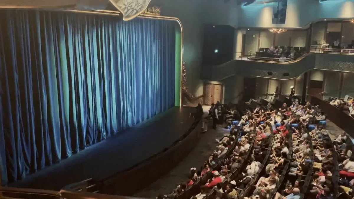A theater with a blue curtain drawn across the stage is filled with an audience seated in anticipation. The elegant interior features ornate balconies and soft lighting, creating a warm and inviting atmosphere.