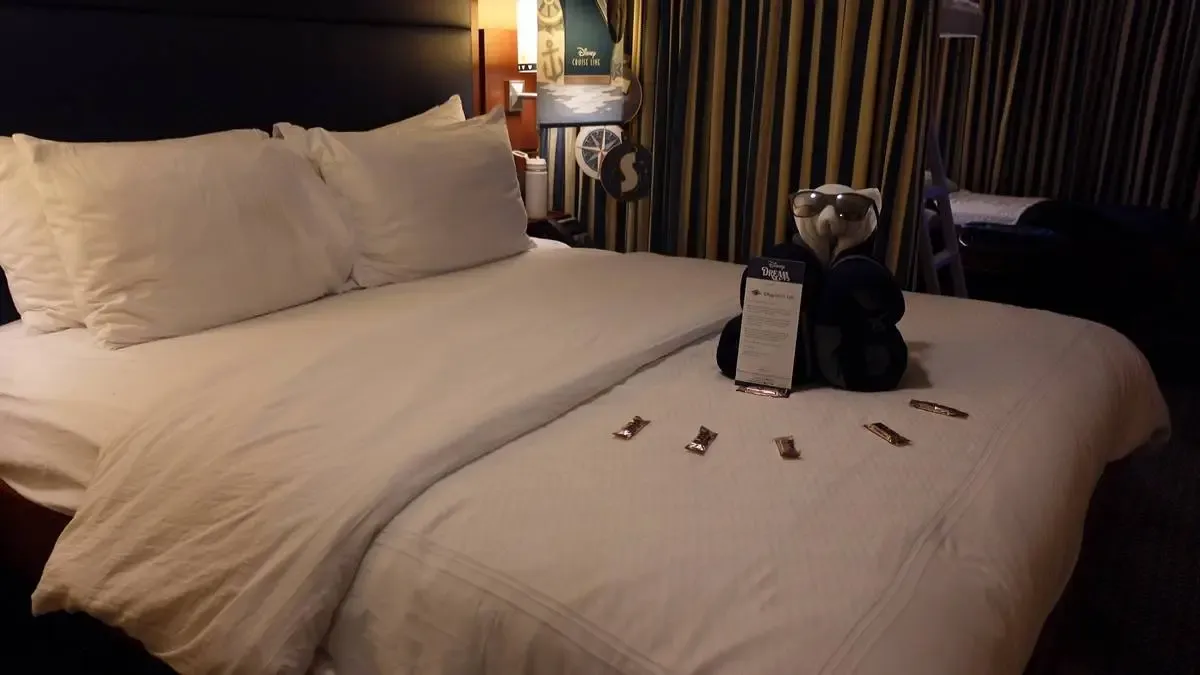 A neatly made hotel bed with crisp white linens and pillows is adorned with a towel animal wearing sunglasses. A card is placed in front of the towel animal, and several small chocolates are arranged on the bedspread. The room has striped curtains and a cozy ambiance with a warm lamp in the background.