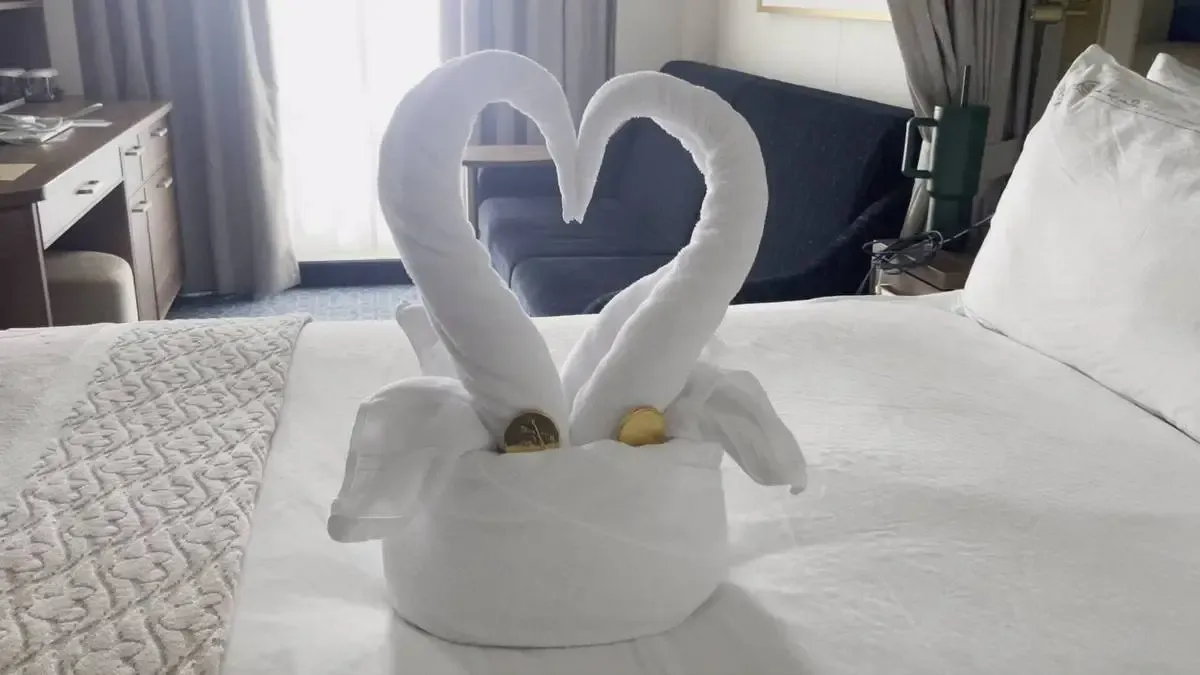 Two white towel swans form a heart shape on a neatly made bed, with a couple of gold-wrapped chocolates nestled at their base, in a cozy, well-lit room.