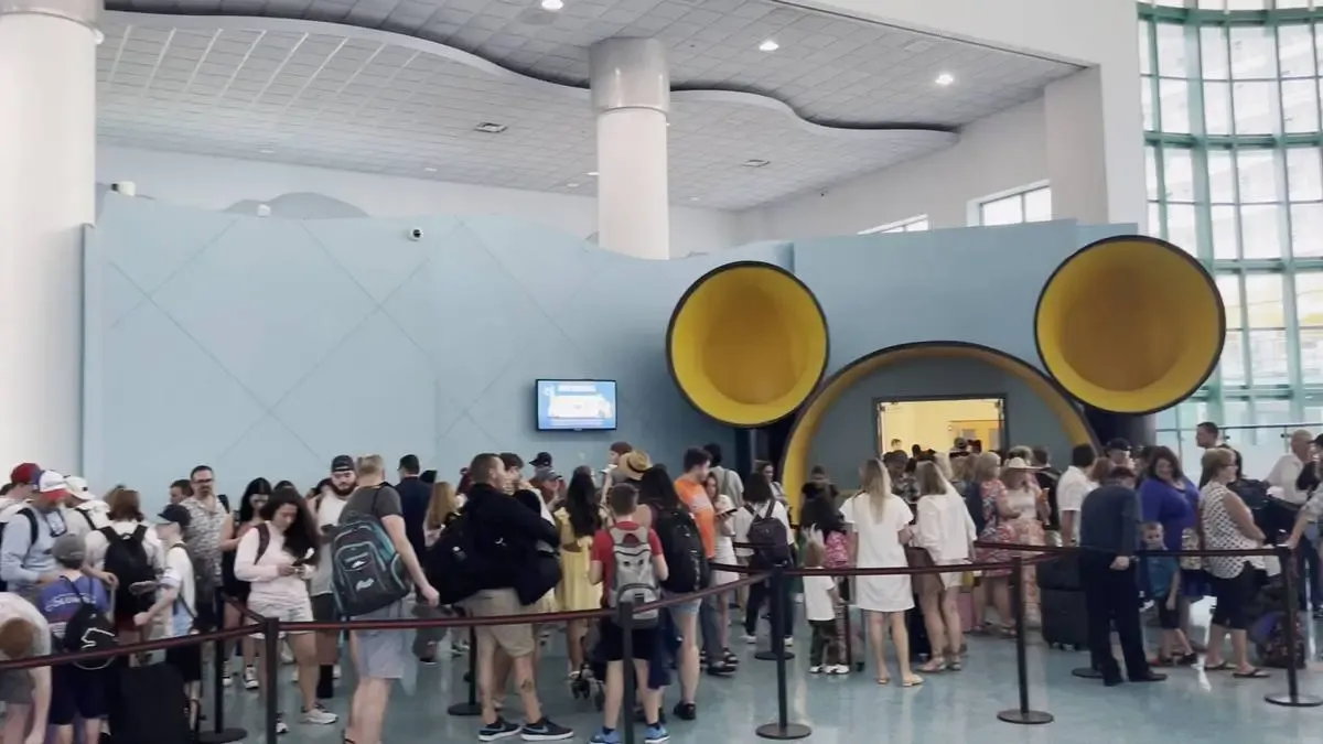 A large group of people, some with backpacks, stand in line inside a spacious building with high ceilings. They are waiting near a blue wall featuring two large, yellow circular openings resembling Mickey Mouse ears, creating a playful atmosphere.