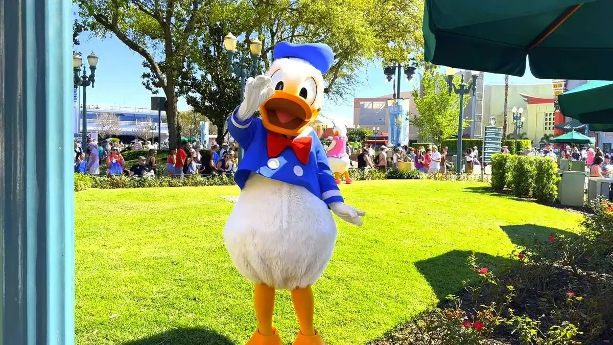 Donald Duck, dressed in his classic blue sailor outfit with a red bow tie, waves cheerfully on a sunny day at a theme park. The lush green lawn and bustling crowd in the background add to the lively atmosphere.