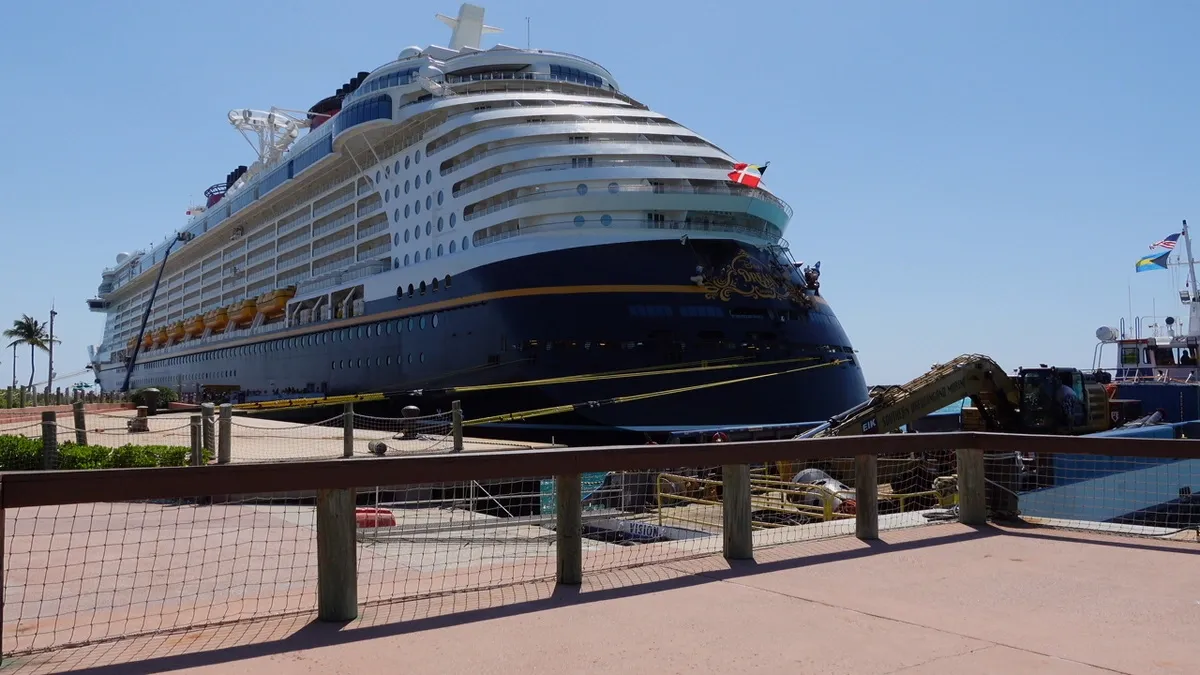 A large cruise ship is docked at a sunny port, featuring a sleek, dark blue hull with white upper decks. Yellow lifeboats are visible along the side, and flags flutter at the bow. A few palm trees are seen in the background, adding to the tropical setting.
