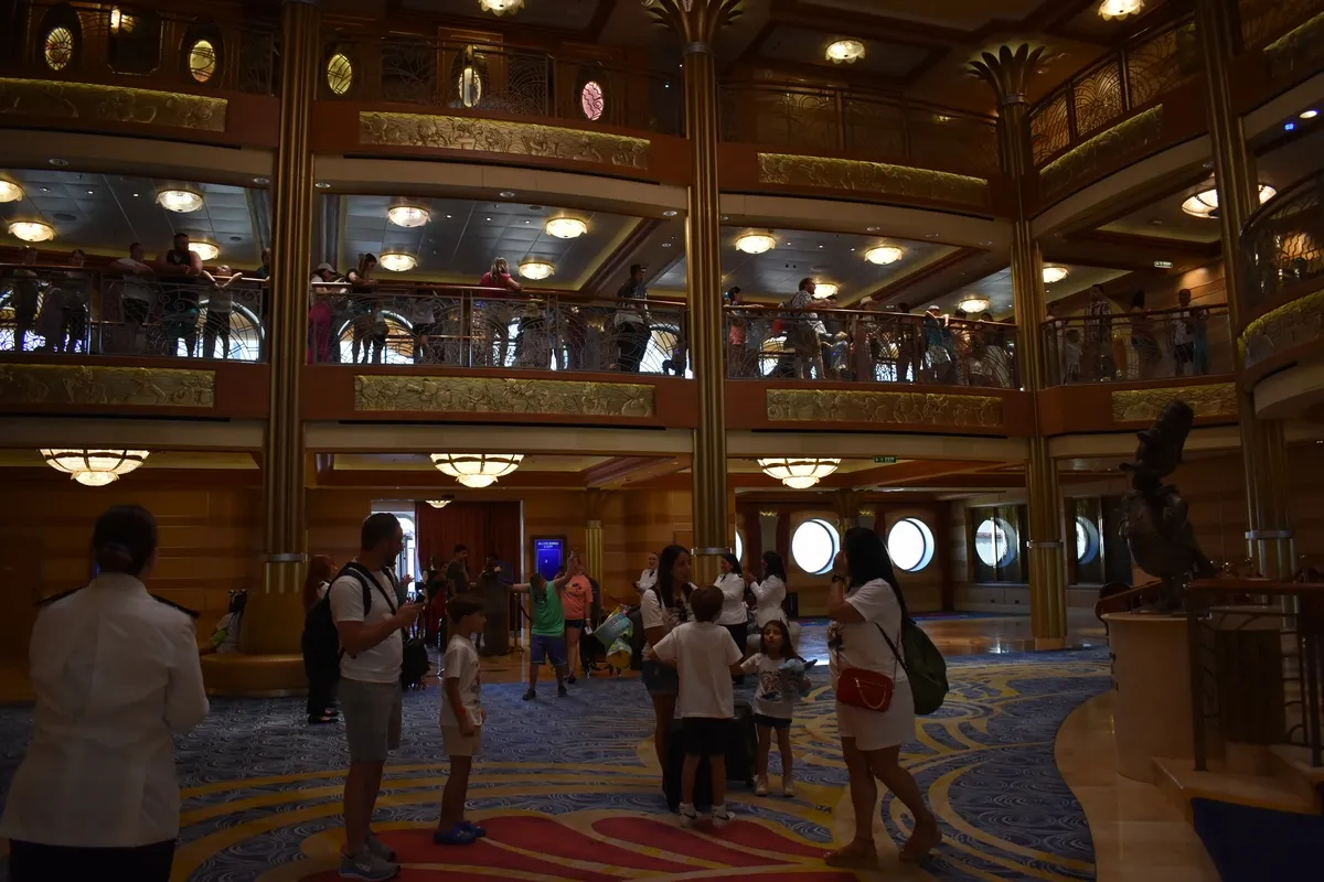 People gather in a spacious, elegant cruise ship atrium with two levels, featuring ornate railings and warm lighting. The carpet has a blue and gold pattern, and a statue is visible on the right. Circular windows and decorative ceiling lights add to the luxurious atmosphere.