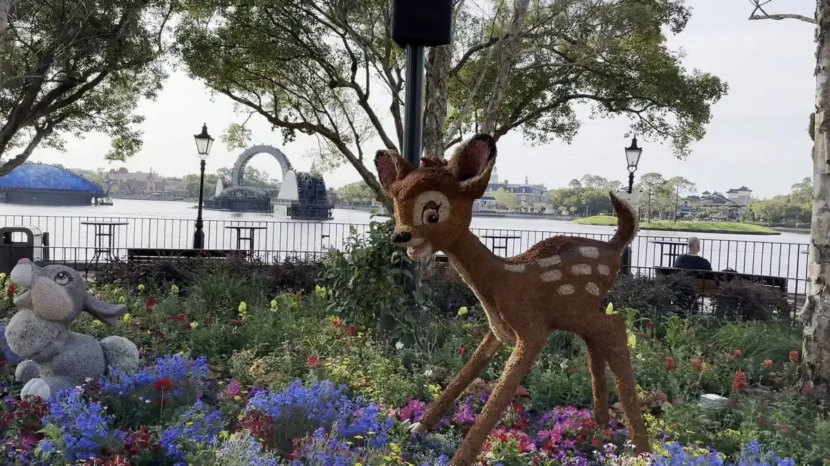 A sculpture of Bambi standing amidst vibrant flowers, with a gray bunny figure nearby. The scene is set in a park by a lake, with trees and a distant view of structures across the water under a clear sky.
