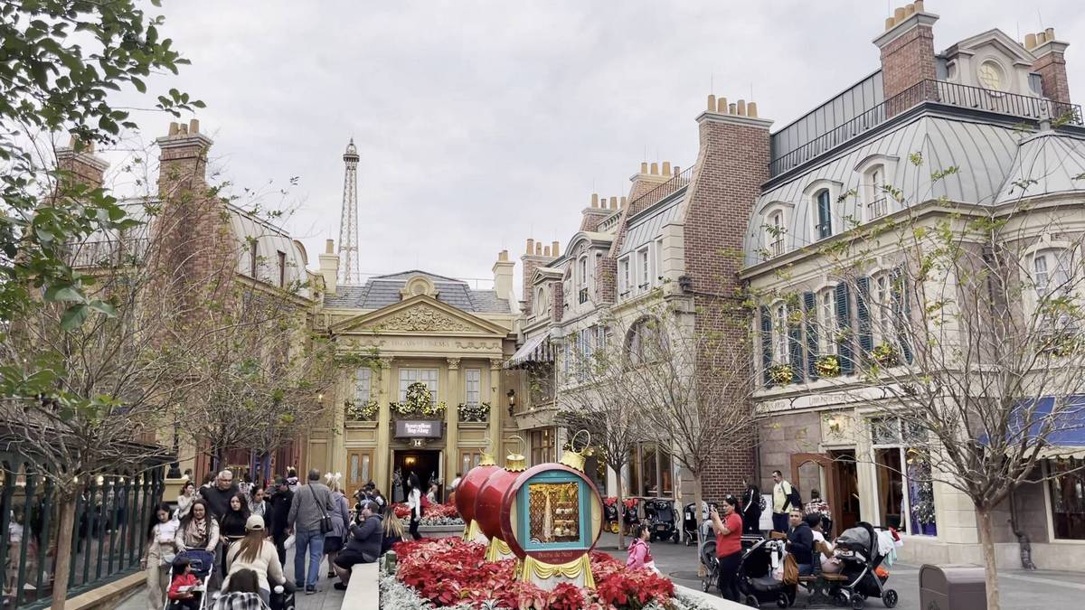 A lively street scene depicts a charming European-style plaza with ornate buildings and a replica of the Eiffel Tower in the background. People stroll along the cobblestone path, some with strollers, surrounded by blooming flowers and bare trees. The atmosphere is bustling and festive.