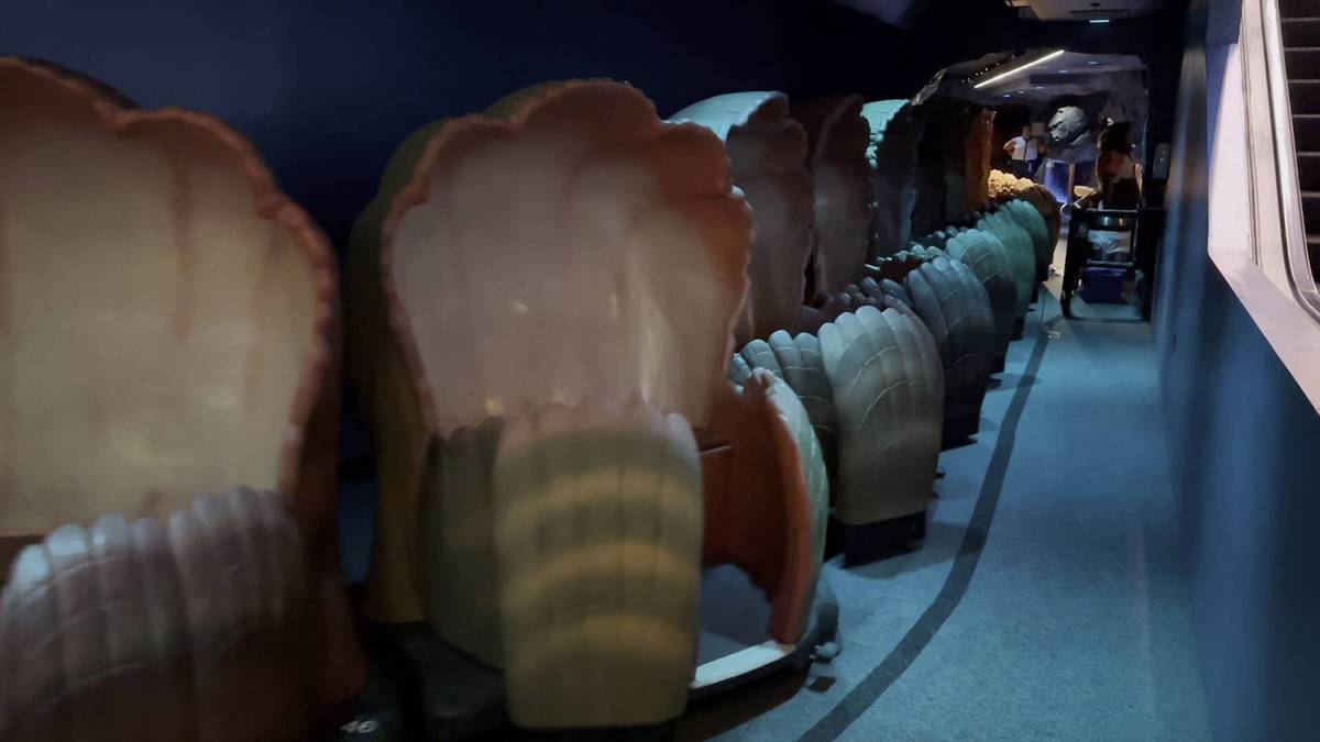 A row of clam-shaped ride vehicles is lined up in a dimly lit indoor setting. The pathway beside them leads toward a softly illuminated area in the distance, where a few people are visible.