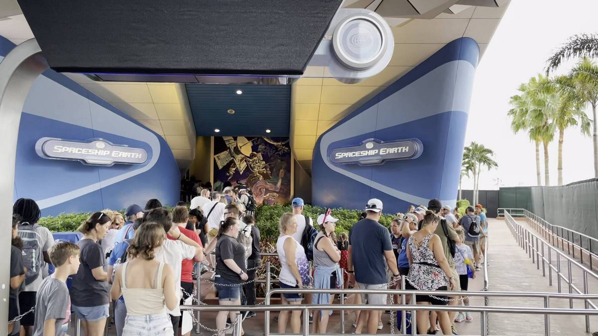 A crowd of people waits in line outside the entrance to Spaceship Earth, marked by a blue and white sign. The setting features modern architecture with angular lines, and palm trees are visible in the background.