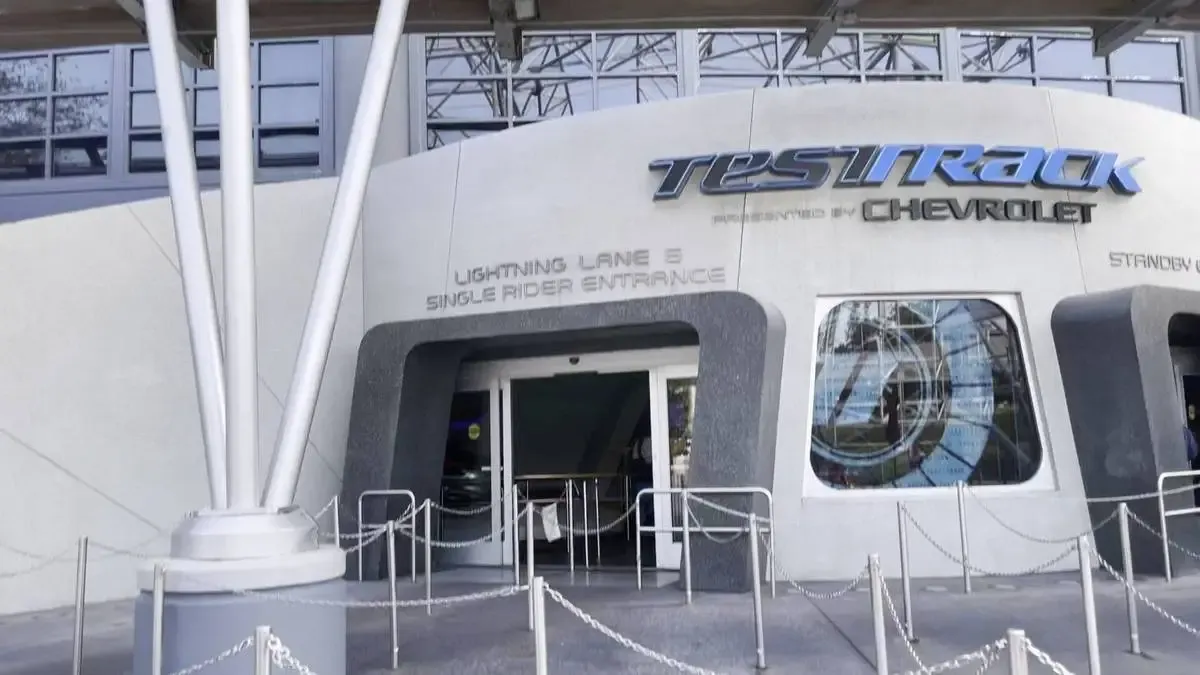 Entrance to the Test Track attraction, featuring a modern facade with a prominent logo above the doorway, and lined queue barriers outside.
