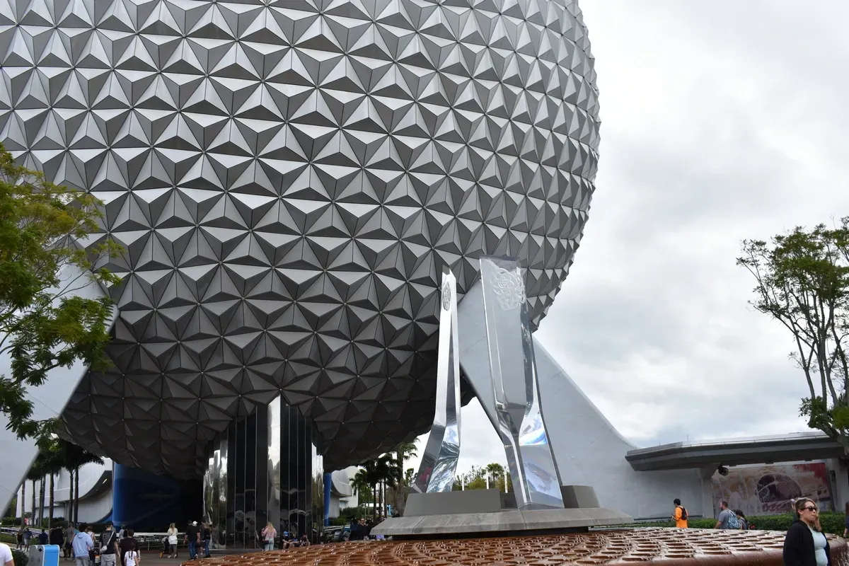 The image shows the iconic geodesic sphere of Spaceship Earth at Epcot, with its triangular silver panels glistening. In the foreground, sleek, modern sculptures stand near the entrance, while visitors walk around the area beneath a cloudy sky.
