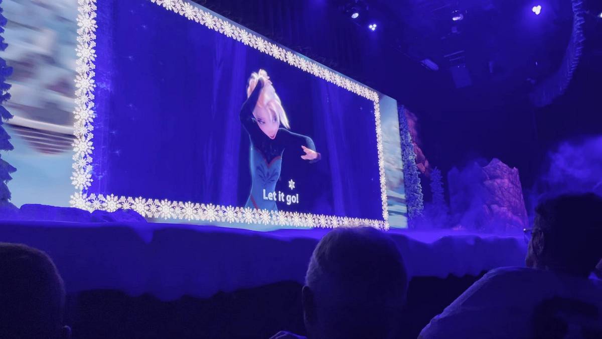 An animated scene from a Frozen is displayed on a large screen in a theater, showing Elsa  with the text "Let it go!" beneath. The screen is framed with a snowflake border, and the audience watches under a blue illuminated atmosphere.