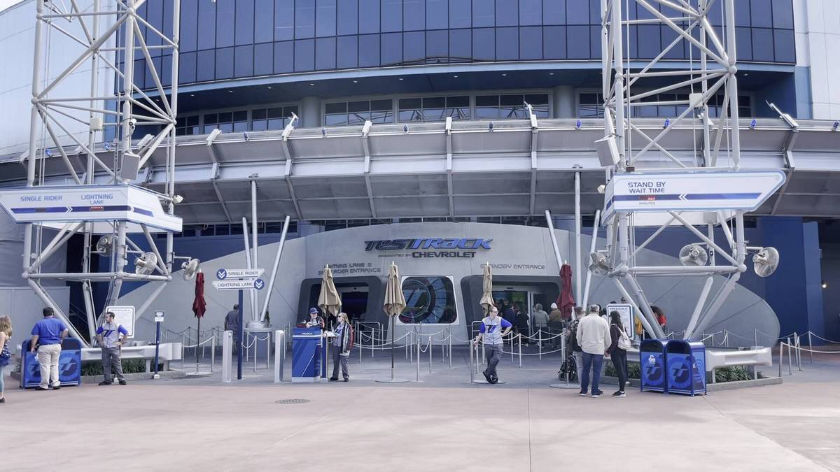 The image shows the entrance to the Test Track attraction. Several people are waiting in line under a futuristic structure with metallic elements. Signs indicate different lanes for single riders and standby wait times.
