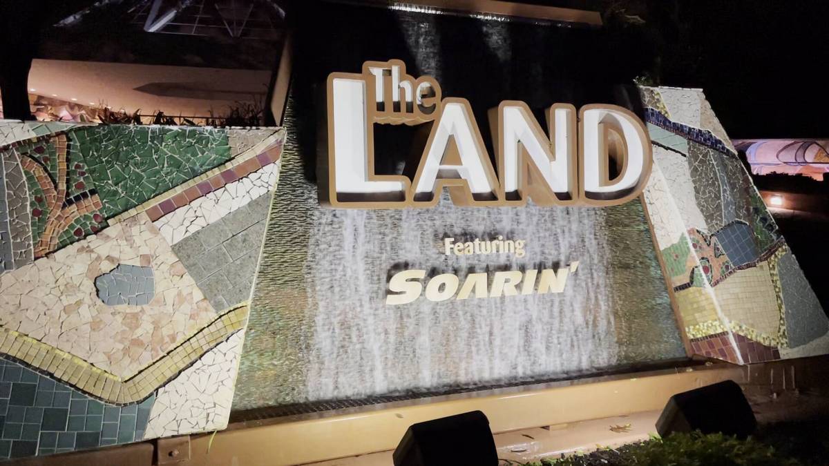 A large sign reading "The Land Featuring Soarin'" is prominently displayed, with a textured mosaic design on either side. The sign is illuminated, highlighting its bold lettering against a backdrop of cascading water.