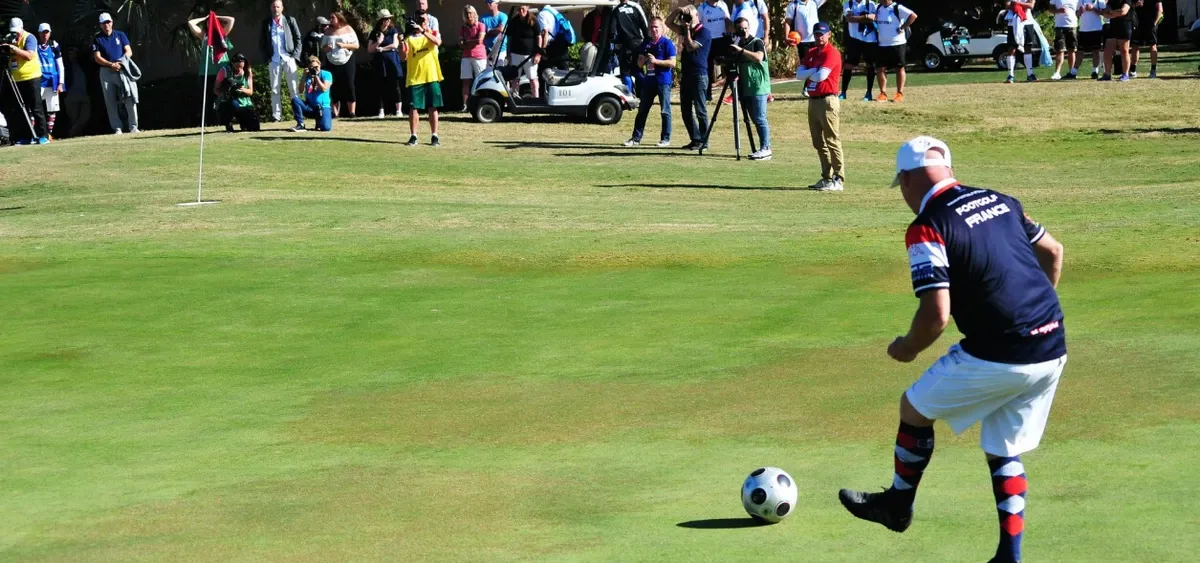 A man wearing a sports jersey and colorful socks is about to kick a soccer ball on a golf course green, with a flag marking the hole nearby. A crowd of spectators watches in the background, some taking photos.