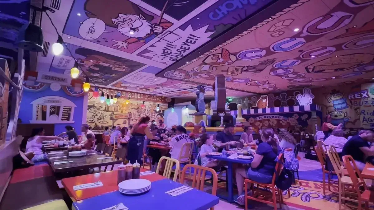 A lively restaurant filled with people dining at colorful tables, featuring a vibrant cartoon-themed decor on the walls and ceiling, with playful illustrations and bright hanging lights enhancing the whimsical atmosphere.