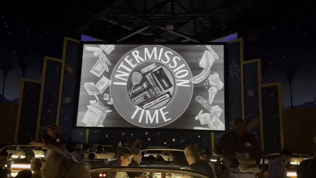 A retro-themed indoor drive-in theater shows a large screen with "Intermission Time" displayed, surrounded by vintage snack illustrations. Below, people sit in car-shaped booths enjoying the ambiance, with dim lighting and a starry backdrop enhancing the nostalgic atmosphere.