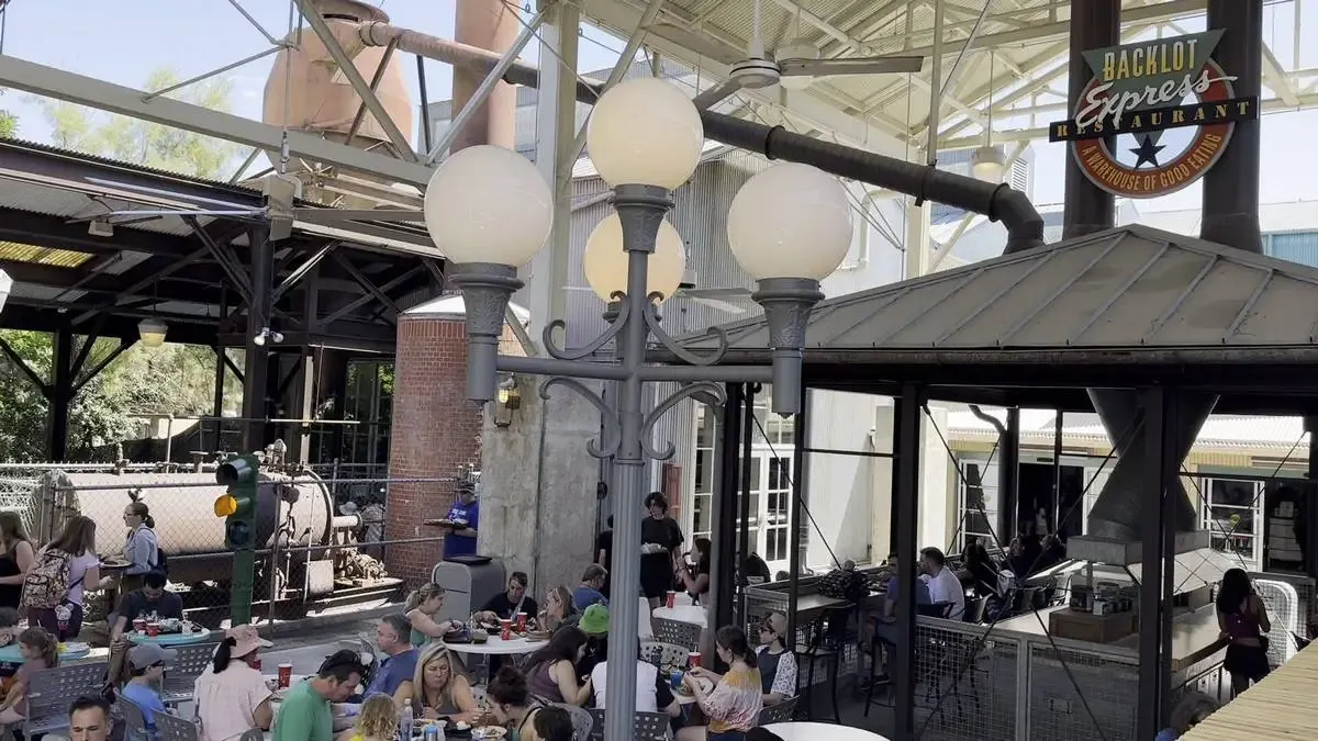 People are dining at outdoor tables under a metal canopy at the Backlot Express Restaurant, with a vintage industrial setting featuring large pipes and a brick structure in the background.