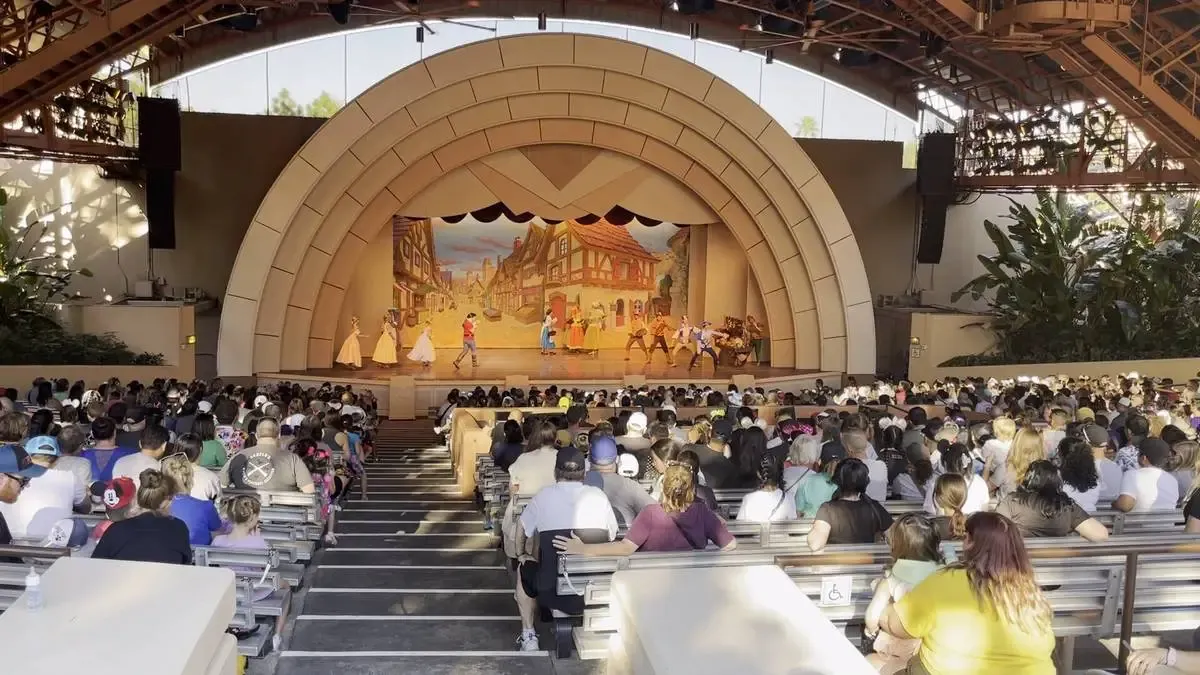A stage performance of Beauty and The Beast unfolds in an outdoor amphitheater. The audience, seated on wooden benches, watches attentively under the large, arching structure that provides shade.