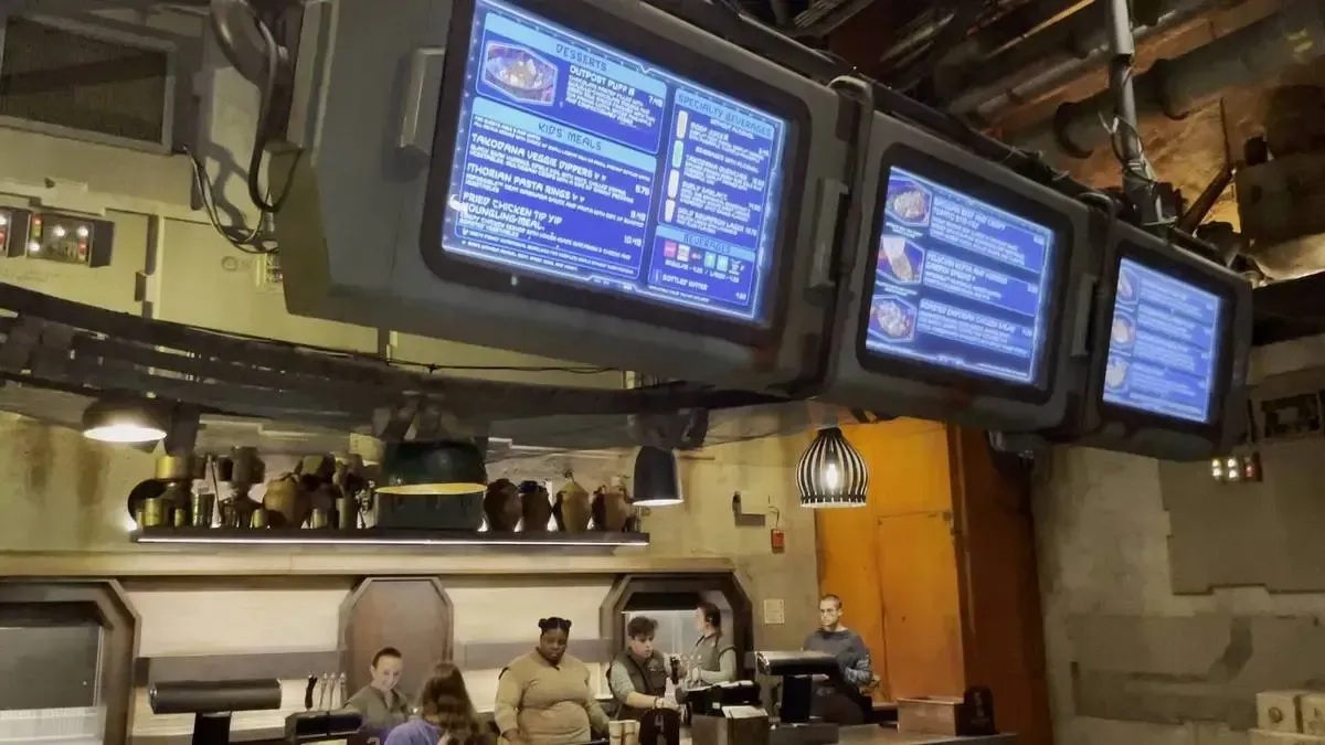 A futuristic restaurant setting features digital menu screens hanging from the ceiling, displaying various food options. Below, staff members assist customers at the counter, with industrial-style decor and warm lighting creating an immersive atmosphere.