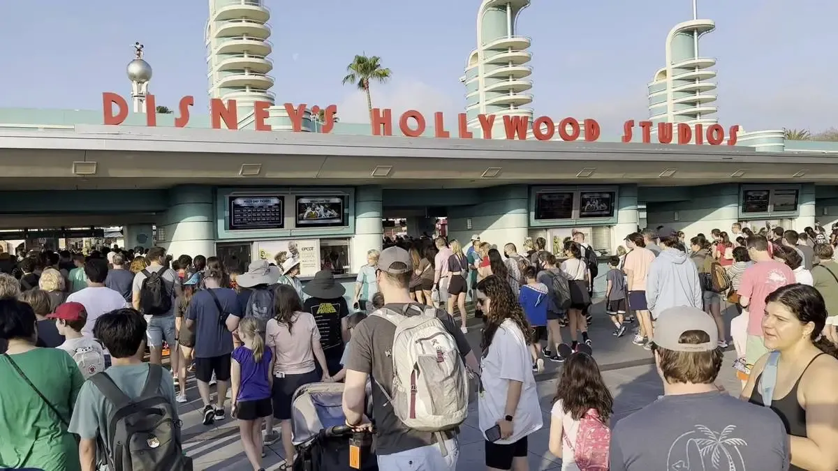 A large crowd gathers at the entrance of Disney's Hollywood Studios. The iconic red signage and art deco-style architecture are visible above the entrance, with palm trees in the background under a clear sky.