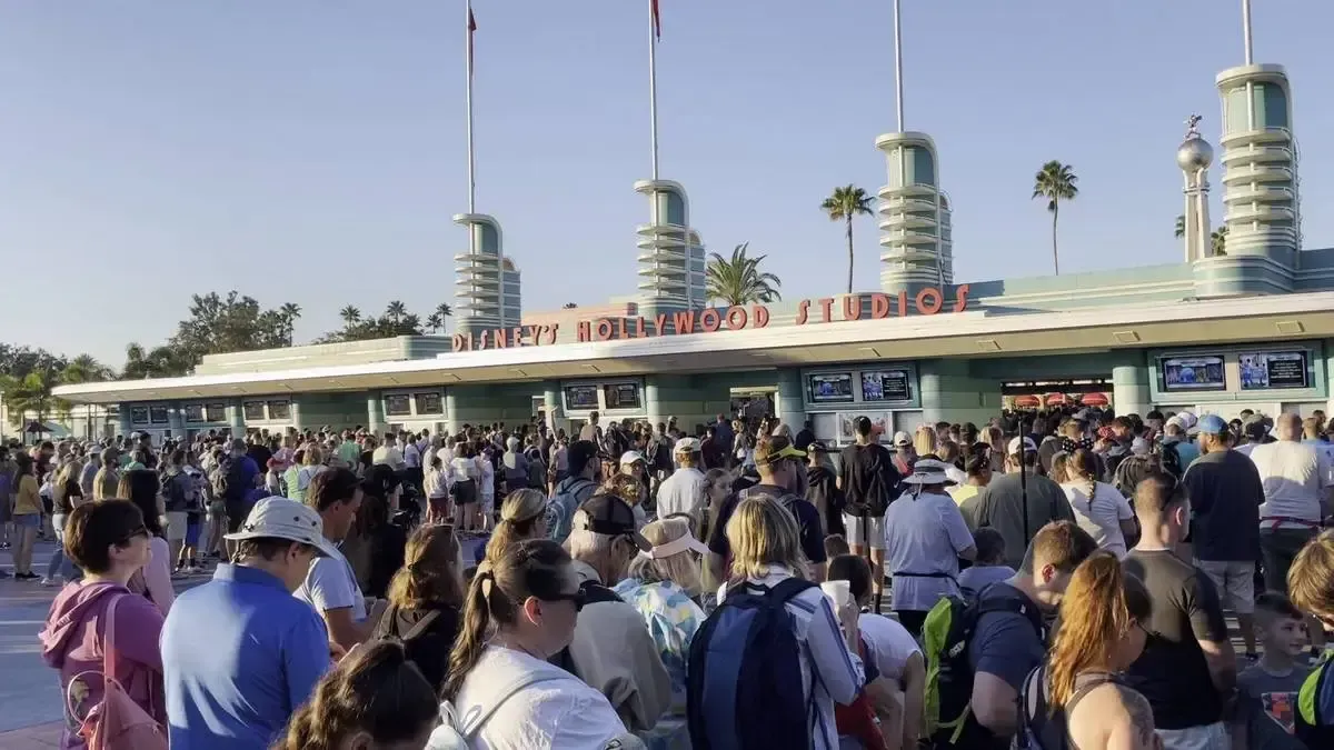 A large crowd of people gathered outside the entrance of Disney's Hollywood Studios, with the art deco-style entrance and palm trees visible in the background under a clear blue sky.