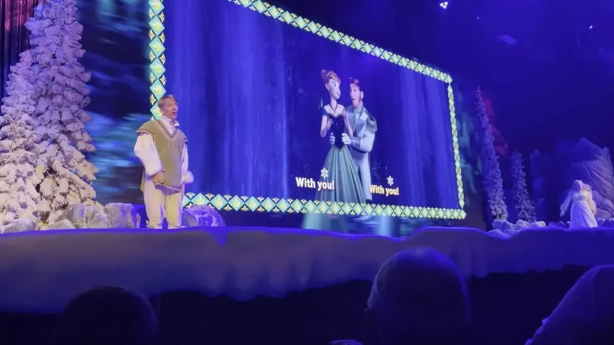 A performer in a winter costume stands on a stage covered in snow beside a large screen displaying an animated scene from "Frozen," featuring two characters dancing. The background is adorned with snowy trees, and the scene is illuminated by blue stage lighting.