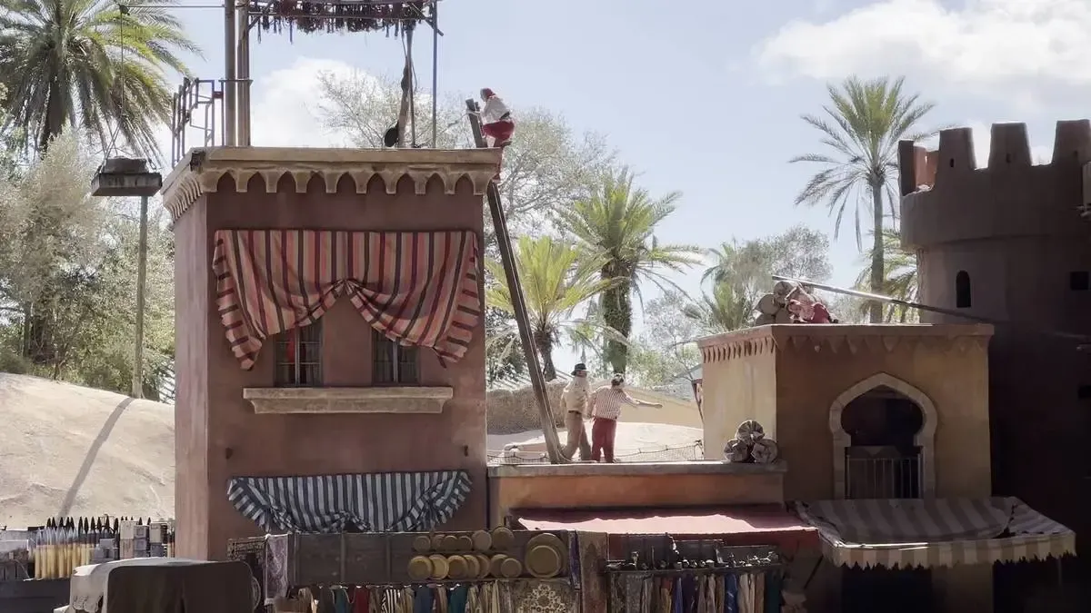 A lively scene depicts performers on a rooftop set designed to resemble an exotic marketplace, with striped awnings and vibrant textiles below. Palm trees and a sandy landscape form the backdrop, while a person climbs a ladder on the left, adding to the sense of adventure.