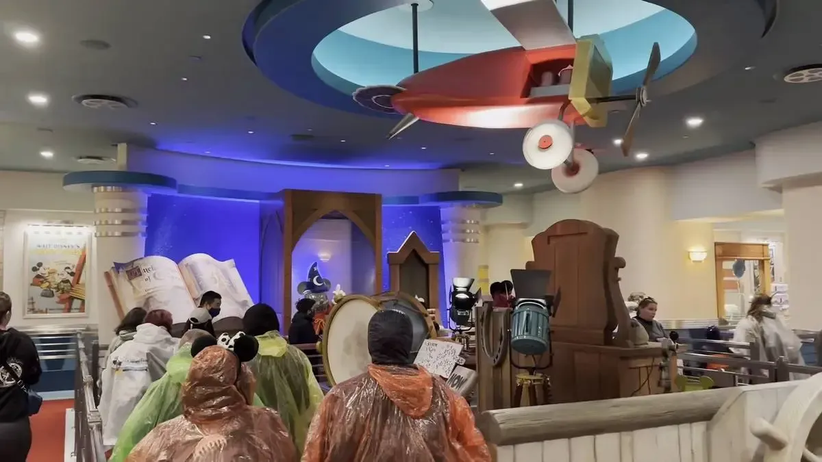 A group of people wearing colorful rain ponchos and Mickey Mouse ears stand in line inside a themed attraction. The room features whimsical decor, including a large open book, and a model airplane hanging from the ceiling.