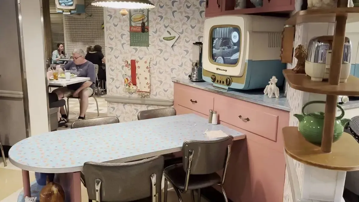 A retro-themed dining area features a vintage television on a pink counter, surrounded by patterned wallpaper and a blue, oval table with metal chairs. A green teapot and other vintage decor adorn the shelves, while people are dining at tables in the background.
