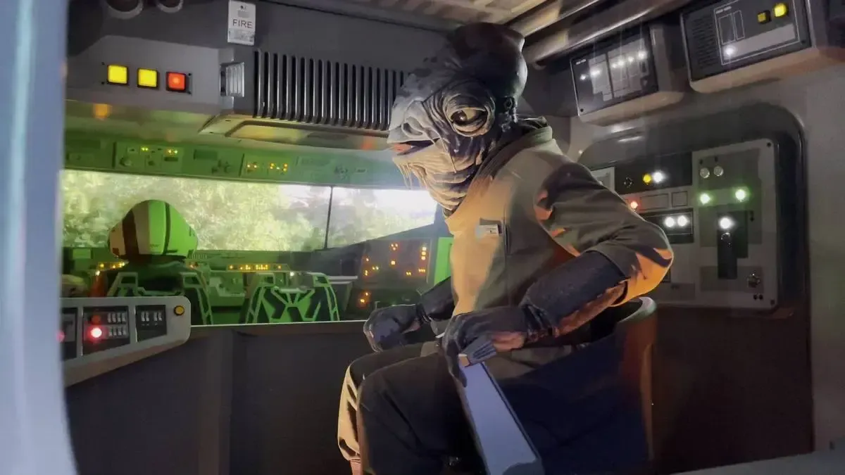An alien character in a brown uniform sits in a futuristic control room filled with glowing buttons and screens, creating a sci-fi atmosphere.