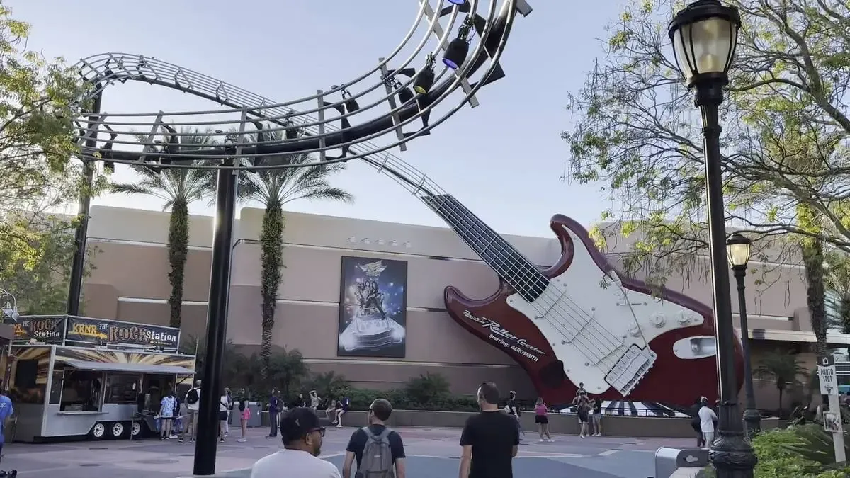 A large, red electric guitar sculpture is integrated into a roller coaster track at an amusement park, with people walking nearby and palm trees in the background.