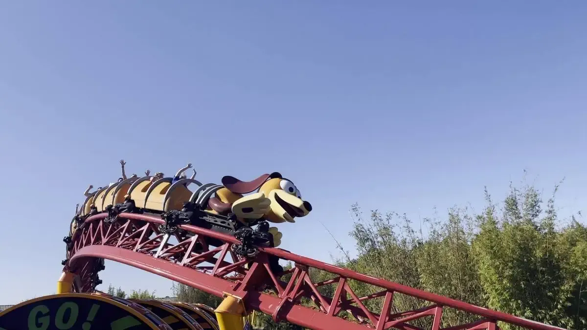 A colorful roller coaster shaped like a playful dog winds along a red track, with passengers raising their arms in excitement under a clear blue sky.