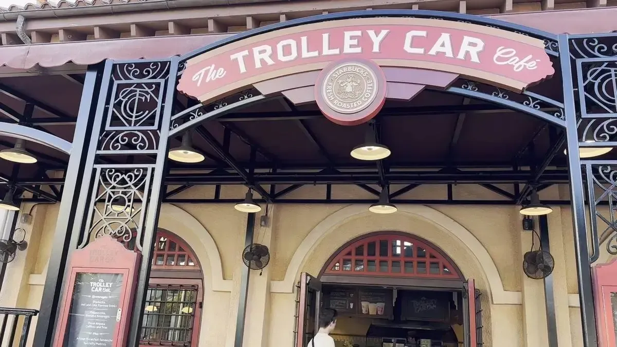 The entrance of The Trolley Car Café, featuring a red and black sign with decorative metalwork and round hanging lights, is set against a beige building facade.