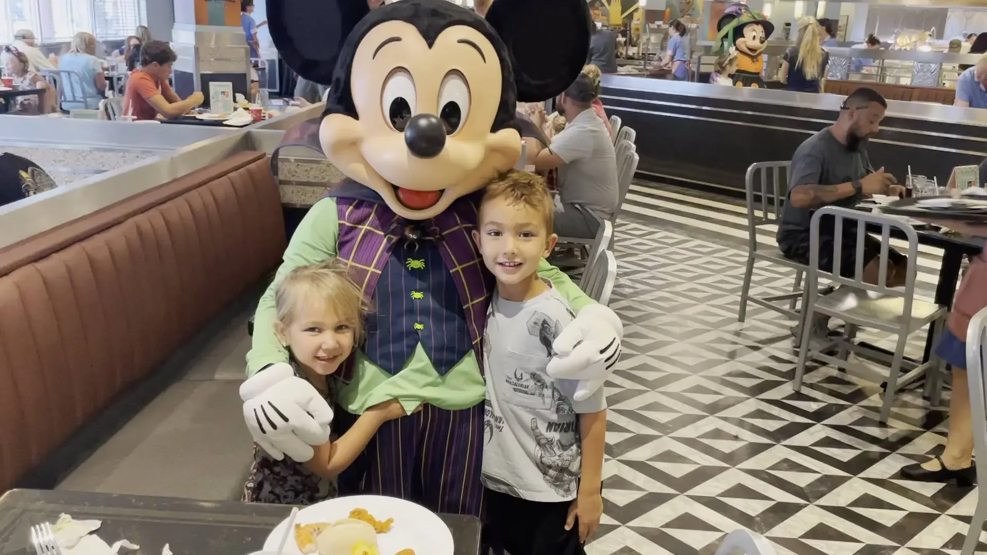 Mickey Mouse, dressed in a colorful costume with a green shirt and purple vest, poses with two smiling children in a lively restaurant setting, with patterned floors and diners in the background.