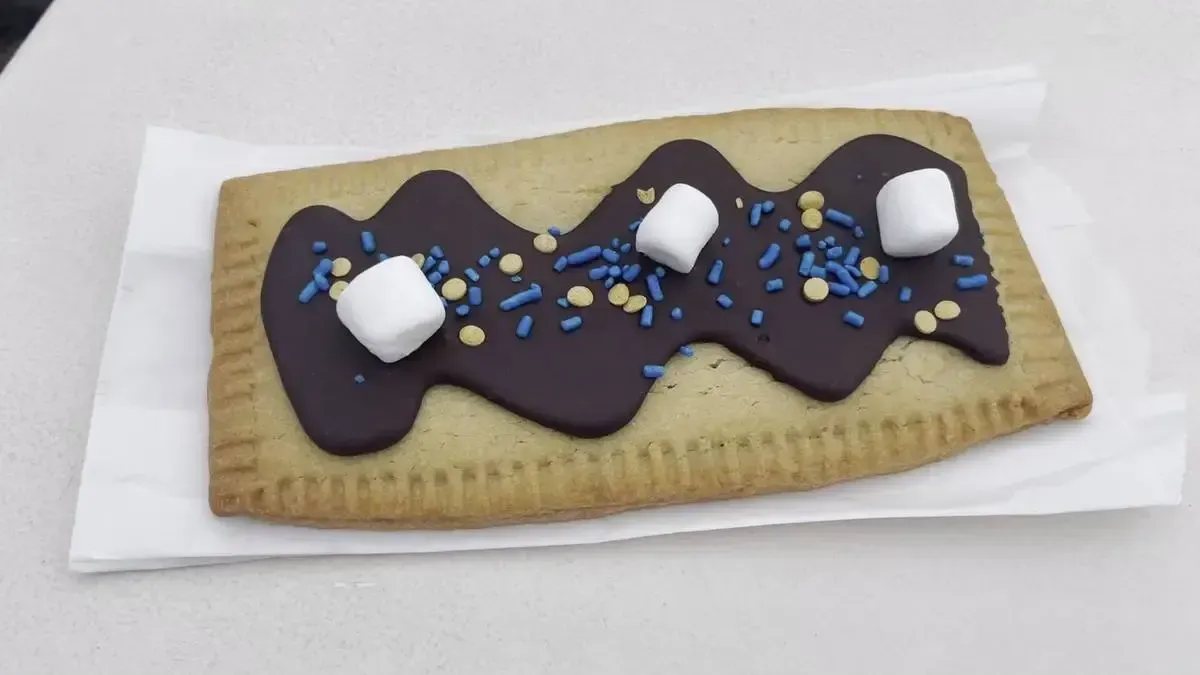 A rectangular pastry topped with a wavy layer of chocolate icing, sprinkled with blue and yellow sprinkles, and adorned with three small marshmallows, is placed on a white napkin.