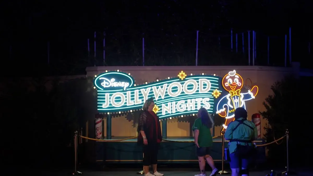 A neon sign glows with "Disney Jollywood Nights" in bright white and blue, featuring a cheerful gingerbread man illustration. Three people stand in front, enjoying the festive atmosphere at night.