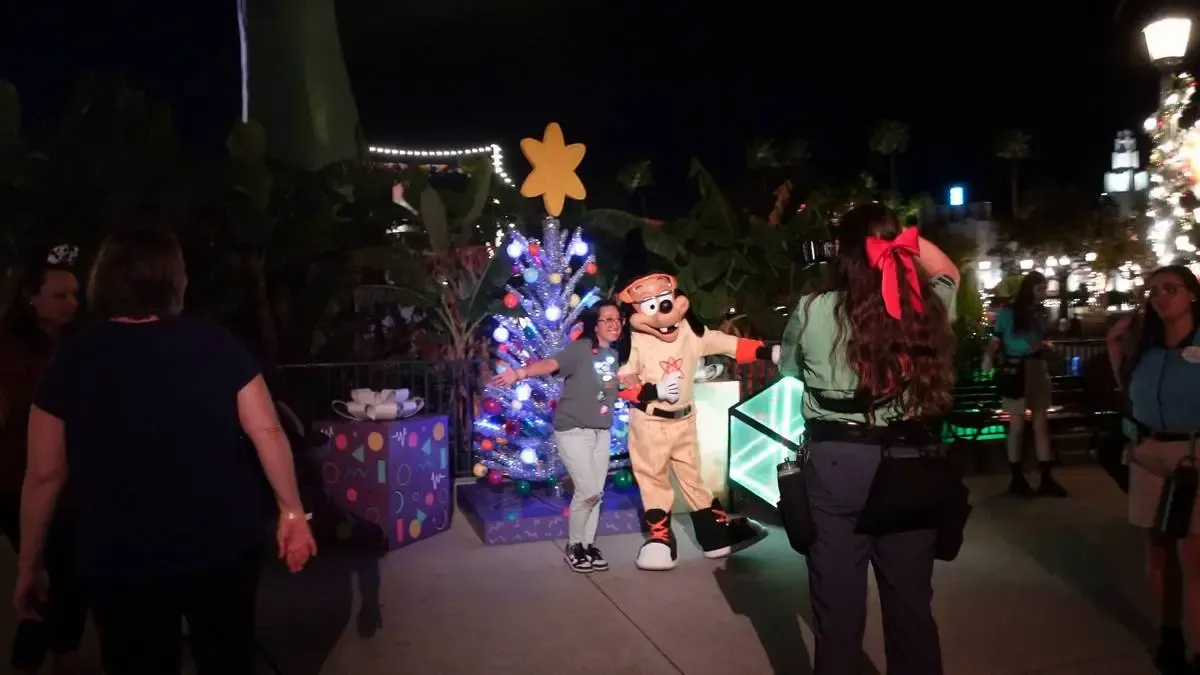 A person poses with a character in front of a festive Christmas tree adorned with colorful lights and ornaments. Several people are around, some taking photos, in a nighttime setting with decorative lights in the background.
