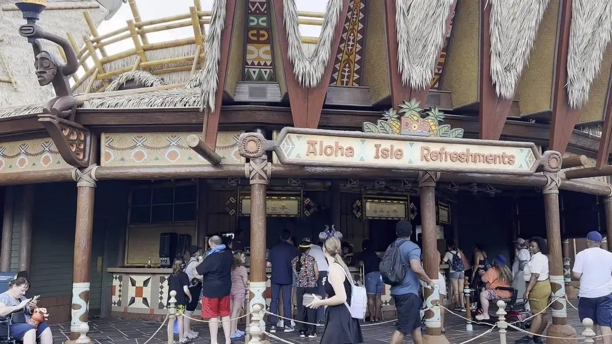 People gather in line at Aloha Isle Refreshments, a tropical-themed stand with a thatched roof and decorative carvings, set in an amusement park.